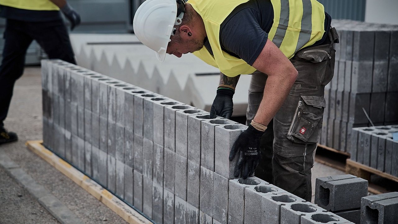 Foto, Bauarbeiter in gelber Sicherheitsweste und weißem Helm beugt sich über eine Reihe Betonsteine, im Hintergrund ein weiterer Arbeiter.