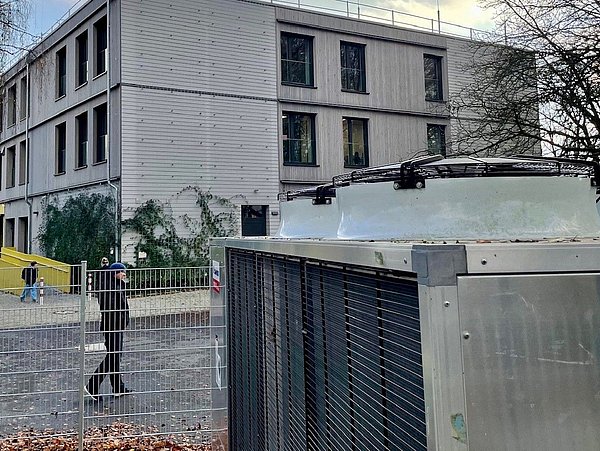 Foto, Technische Anlage eingezäunt vor grauem Gebäude, im Hintergrund laufen zwei Menschen vorbei über das Gelände, Himmel grau. 