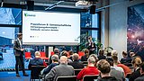 Foto, Zwei Personen stehen vor Publikum in einem Seminarraum, eine präsentiert vor einer Leinwand mit Text zu gemeinschaftlichen Versorgungskonzepten.