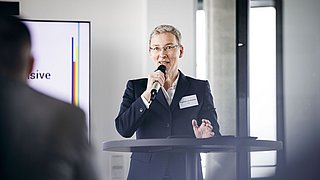 Foto, Frau im dunklen Anzug hält Mikrofon und spricht an einem Stehtisch bei einer Präsentation