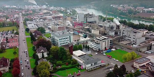 Luftaufnahme eines Industriegebiets mit Fabrikgebäuden, Schornsteinen und angrenzenden Straßen neben einem Fluss, aufsteigendem Rauch von industrieller Abwärme.