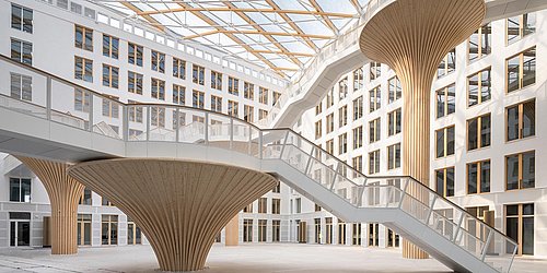 Foto, weitläufiges Foyer mit aus Holz gefertigten Säulen und Treppen. Rings um das Foyer befinden sich Wände mit Fenstern auf mehreren Stockwerken. 