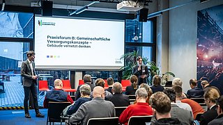 Foto, Zwei Personen stehen vor Publikum in einem Seminarraum, eine präsentiert vor einer Leinwand mit Text zu gemeinschaftlichen Versorgungskonzepten.