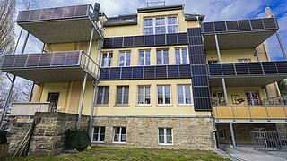Foto, Mehrstöckiges Wohngebäude mit gelber Fassade und mehreren Balkonen, die mit Solarpaneelen bedeckt sind