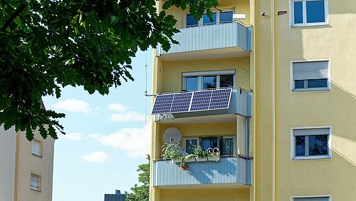 Foto, ein gelbes Wohnhaus mit Balkonen mit Sonnenkollektoren und Bepflanzung, umrahmt von üppigen grünen Blättern unter einem blauen Himmel.