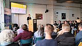 Foto, Person steht an einem Rednerpult vor Publikum in einem Raum mit sichtbarer Ziegelwand und einem Bildschirm mit Präsentationsfolien.