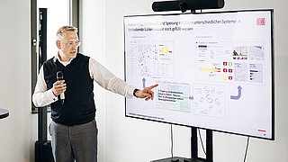 Foto, Mann mit Mikrofon zeigt auf Bildschirm mit Präsentationsfolien und Diagrammen.