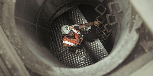 Foto, ein Arbeiter in Arbeitskleideung steht in einem Schacht und schaut hoch zur Öffnung.