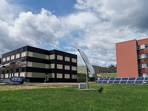 Zwei Gebäude auf einer Wiese: links ein dreigeschossiges, schwarz-grünes Gebäude mit einem Flugzeug auf dem Dach, rechts ein rotes, dreigeschossiges Gebäude mit Balkonen. Im Vordergrund stehen Solarpanels.