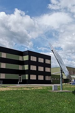 Foto, zwei Gebäude auf einer Wiese: links ein dreigeschossiges, schwarz-grünes Gebäude mit einem Flugzeug auf dem Dach, rechts ein rotes, dreigeschossiges Gebäude mit Balkonen. Im Vordergrund stehen Solarpanels.