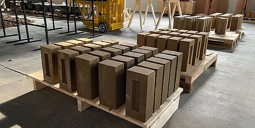 Foto, gepresste Lehmsteine auf zwei Holzpaletten gestapelt.