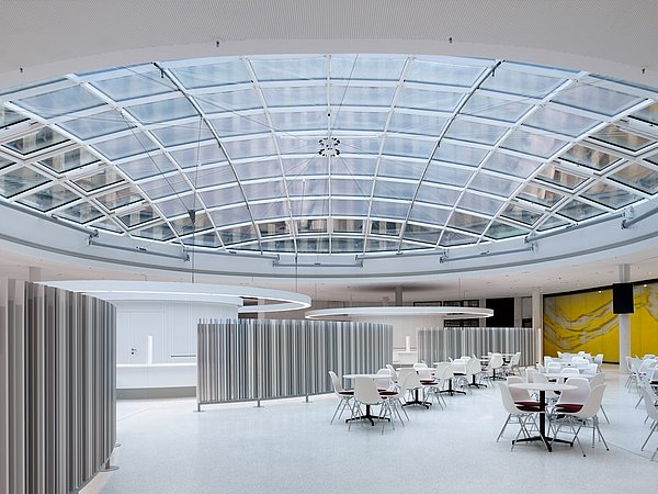 Foto, großer, heller Innenraum mit mehreren kleinen Tischen und Stühlen unter einem kuppelähnlichen Dach aus Glas.