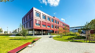 Foto, Außenansicht eines roten Gebäudes mit zwei Stockwerken. Im Vordergrund befinden sich ein Weg und Grünflächen.