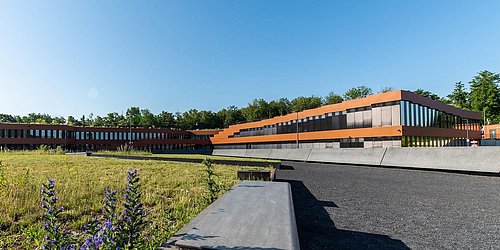 Foto, Modernes Bürogebäude der RAG-Stiftung mit großen Glasfenstern, umgeben von Wiesen und Bäumen unter klarem Himmel