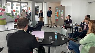 Foto, Mehrere Personen sitzen und hören einer stehenden Frau zu, die in einem modernen Büro mit Glaswänden und bunten Notizen an der Wand spricht.