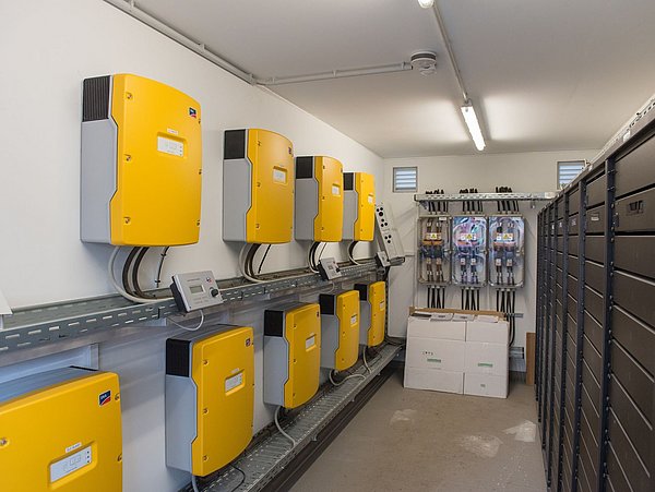 Foto, Technikraum mit acht gelben Wechselrichtern an der Wand und mehreren Stromzählern in einem Gebäude