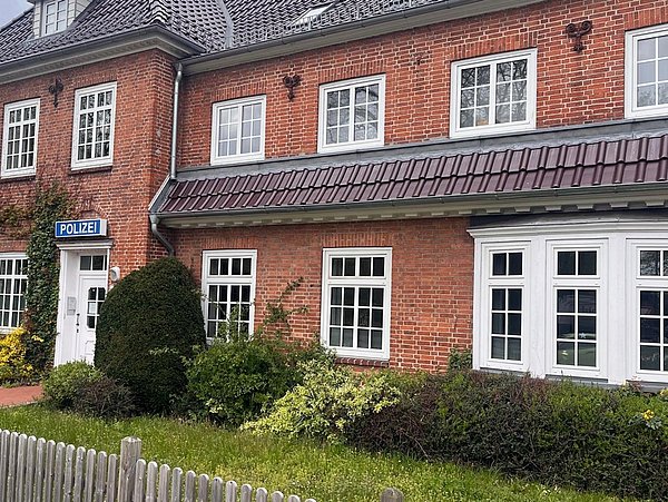 Foto, Zweistöckiges rotes Backsteingebäude mit mehreren weißen Fenstern, einem Schild mit der Aufschrift 'POLIZEI' und einem kleinen Garten mit Sträuchern davor.