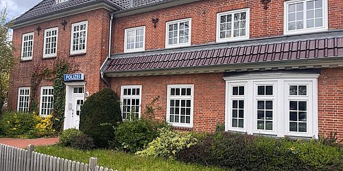 Foto, Zweistöckiges rotes Backsteingebäude mit mehreren weißen Fenstern, einem Schild mit der Aufschrift 'POLIZEI' und einem kleinen Garten mit Sträuchern davor.