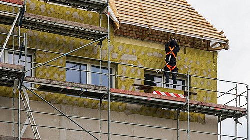Foto, Bauarbeiter mit Sicherheitsgeschirr steht auf einem Gerüst vor einer teilweise mit Dämmmaterial verkleideten Hausfassade unter einem Dach mit sichtbaren Dachlatten.