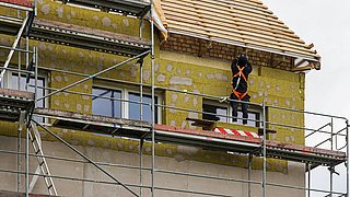 Foto, Bauarbeiter mit Sicherheitsgeschirr steht auf einem Gerüst vor einer teilweise mit Dämmmaterial verkleideten Hausfassade unter einem Dach mit sichtbaren Dachlatten.