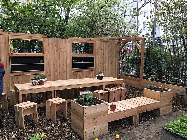 Foto, Tische, Stühle und freistehende Wände mit Fenstern im Außenbereich. Alles ist aus Holz gefertigt und ergibt zusammen ein sogenanntes "Grünes Klassenzimmer". Zur Website des BAUFACHFRAU Berlin e.V.