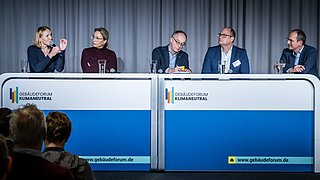 Foto, fünf Personen sitzen an zwei nebeneinanderstehenden Tischen mit der Aufschrift "Gebäudeforum Klimaneutral", vor ihnen stehen Gläser mit Wasser, im Vordergrund Publikum.