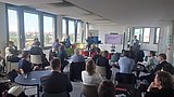 Foto, Konferenzraum mit mehreren Personen, die einem Vortrag folgen, zwei Vortragende vor einem Bildschirm mit Präsentation