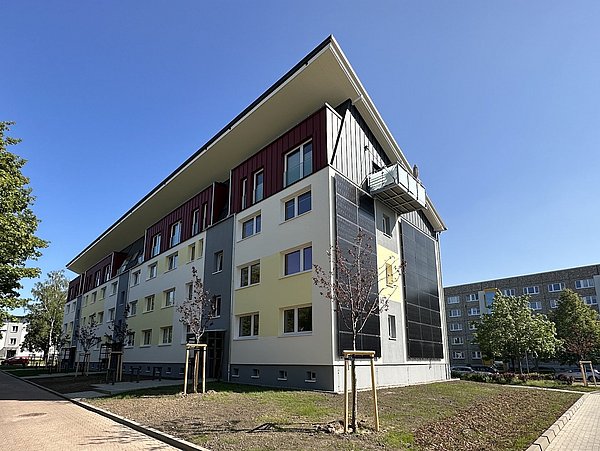 Mehrstöckiges Wohngebäude nach der Sanierung mit gelb-weißem Putz und dunklen Fassadenelementen, umgeben von jungen Bäumen und Gehwegen unter blauem Himmel.