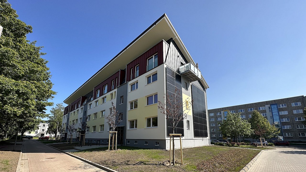 Mehrstöckiges Wohngebäude nach der Sanierung mit gelb-weißem Putz und dunklen Fassadenelementen, umgeben von jungen Bäumen und Gehwegen unter blauem Himmel.
