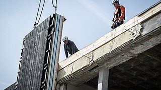 Foto, Zwei Bauarbeiter auf einer Baustelle heben mit einem Kran eine große Wandplatte an einem Gebäude