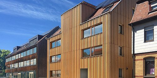 Foto, Straßenansicht mehrerer Gebäude. Im Mittelpunkt stehen zwei mit Holz verkleidete, mehrstöckige Gebäude.