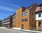 Foto, Straßenansicht mehrerer Gebäude. Im Mittelpunkt stehen zwei mit Holz verkleidete, mehrstöckige Gebäude.