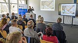 Foto, Mann hält Vortrag vor Publikum in hellem Raum mit zwei Bildschirmen, die ein Dachstuhlfoto zeigen