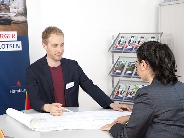 Foto, zwei Personen sitzen sich gegenüber an einem Tisch, auf dem ein Planungsbogen liegt. Im Hintergrund steht ein Regal mit Broschüren. 