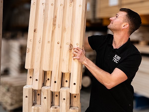 Foto, Mann in schwarzem Poloshirt hält mehrere längliche, helle Holzelemente mit Bohrlöchern in einer Lagerhalle.