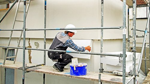Foto, ein Bauarbeiter kniet auf einem Baugerüst und montiert Dämmplatten an einem Gebäude.