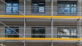 Foto, Fassade eines Gebäudes mit grauen vertikalen Paneelen und schwarzen Fenstern, eingerüstet mit Metallgerüst und gelben Schutzstreifen