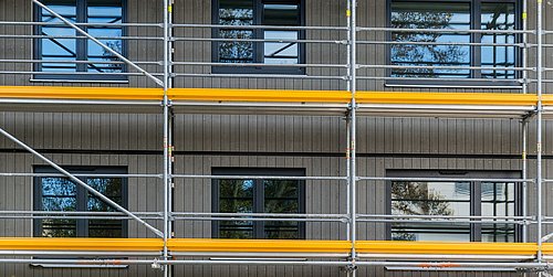 Foto, Fassade eines Gebäudes mit grauen vertikalen Paneelen und schwarzen Fenstern, eingerüstet mit Metallgerüst und gelben Schutzstreifen
