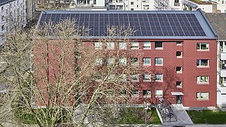 Foto, Mehrstöckiges rotes Wohngebäude mit Solarpaneelen auf dem Dach und kahlen Bäumen im Vordergrund