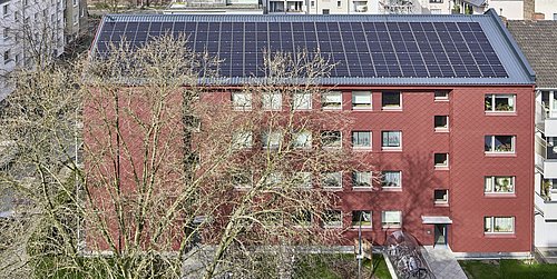 Foto, Mehrstöckiges rotes Wohngebäude mit Solarpaneelen auf dem Dach und kahlen Bäumen im Vordergrund