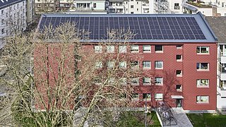 Foto, Mehrstöckiges rotes Wohngebäude mit Solarpaneelen auf dem Dach und kahlen Bäumen im Vordergrund