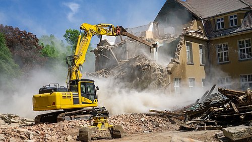Foto, Gelber Bagger reißt ein altes Gebäude mit gelben Wänden und braunem Dach ab, Staub steigt auf