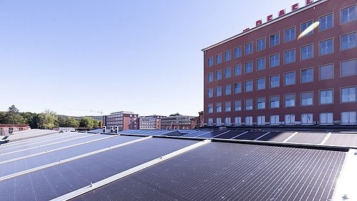 Foto, Dach mit mehreren Solarpaneelen vor einem mehrstöckigen Bürogebäude unter klarem Himmel.