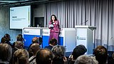 Foto, Frau in pinkem Kostüm hält Notizen und spricht vor Publikum in einem Konferenzraum mit grauen Vorhängen und Präsentationsbildschirm.
