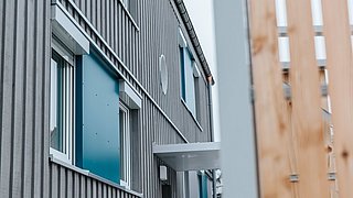 Foto, Bild eines modernen Gebäudes mit grauer vertikaler Verkleidung und blauen Fensterverkleidungen, teilweise verdeckt durch einen Holzzaun.