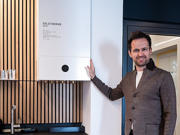 Mann in brauner Strickjacke steht neben weißer Soletherme Mini an Holzlattenwand und berührt das Gerät mit der Hand.