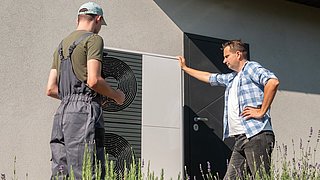 Foto, zwei Männer stehen vor einer Wand mit einer großen Wärmepumpe, einer trägt graue Latzhose und eine Kappe, der andere ein kariertes Hemd und Jeans.