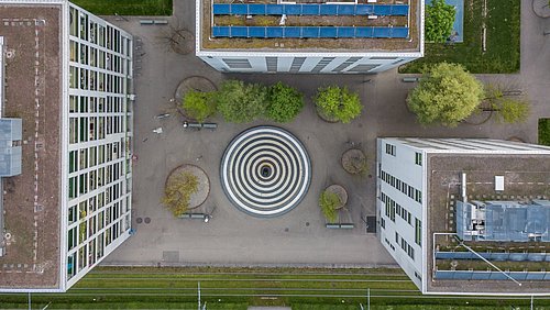 Foto, Luftaufnahme dreierGebäude mit PV-Modulen auf dem Dach, die sich um einen kreisrunden Brunnen gruppieren.