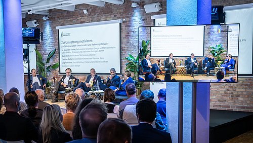 Foto, Podiumsdiskussion bei der Jahrestagung 2024 mit fünf Personen auf der Bühne vor Publikum, Präsentation zeigt Motivationspunkte zur Umsetzung