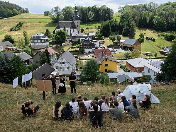 Foto, eine Gruppe sitzt auf einem Hügel, im Hintergrund befindet sich ein kleines Dorf mit einer Kirche